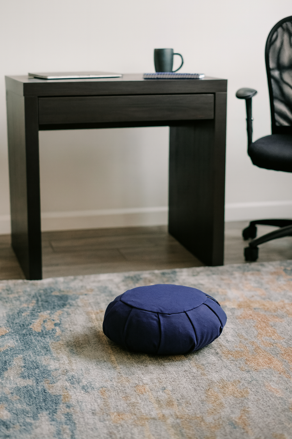 Blue meditation cushion on a patterned rug in front of a black desk with a chair.