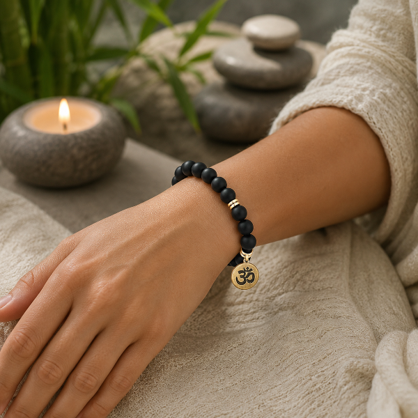 Hand wearing a black beaded bracelet with a gold Om symbol charm, set against a blurred natural background.