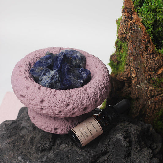 Pink stone bowl with blue crystal, black bottle labeled 'Nurturing' on a rock with moss.