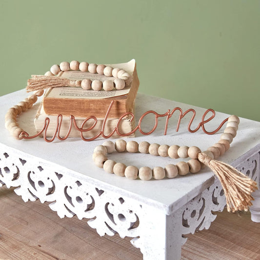 Decorative 'welcome' sign with wooden beads and tassels on a white surface.